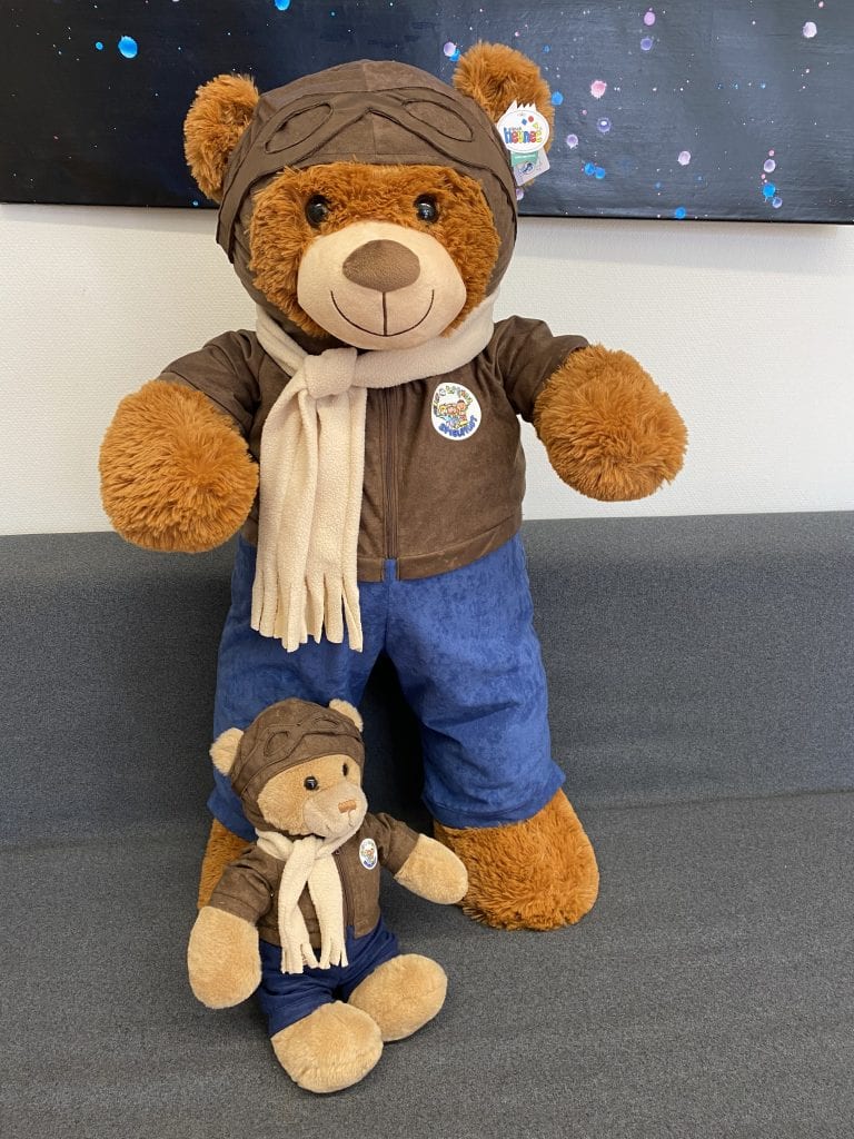Braunes Piloti-Maskottchen aus Plüsch mit brauner Mütze und blauem Outfit steht vor einer farbenfrohen Wand mit blauen und pinken Punkten.