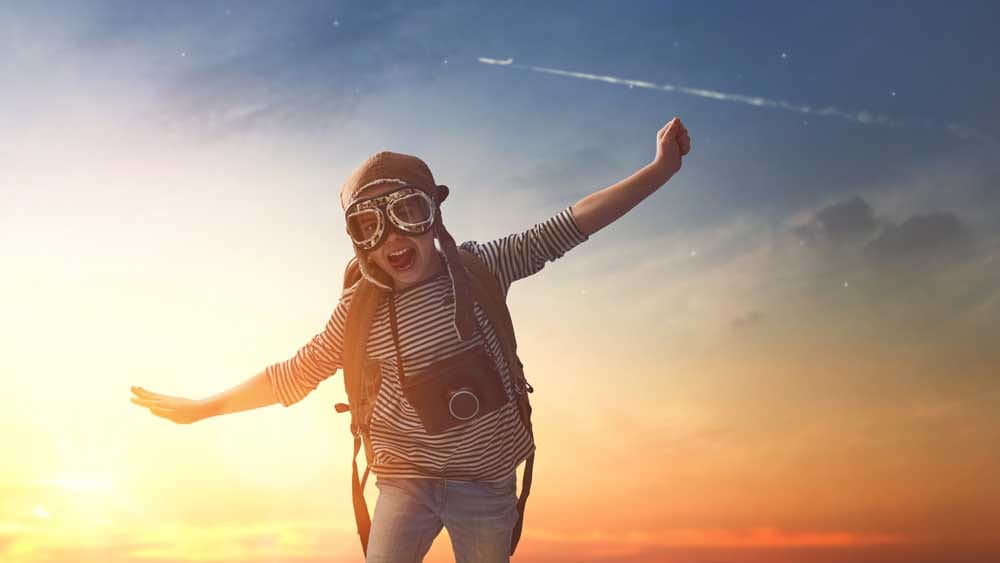 Im goldenen Abendlicht trägt ein Kind eine Aviatorbrille und ein gestreiftes Shirt, Arme weit ausgebreitet vor dem Himmel – wie ein Flieger.