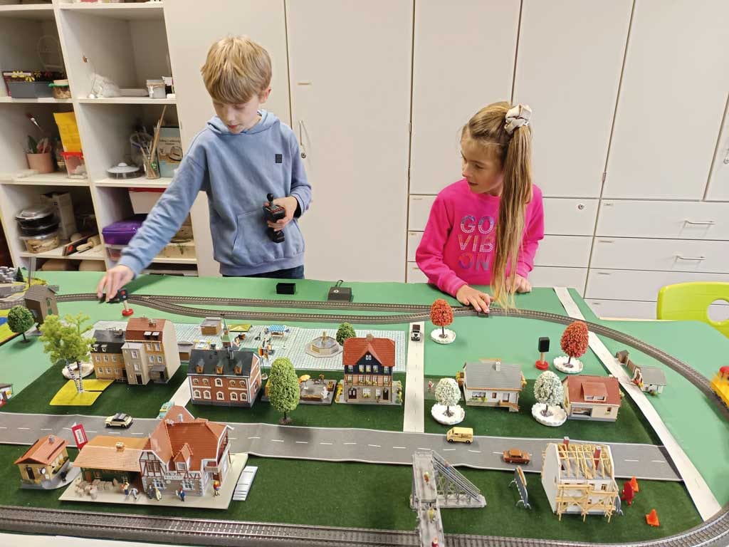 Auf einer grünen Landschaft liegen graue Gleise mit kleinen Häusern in Braun und Ziegeloptik, daneben stehen Bäume und Fahrzeuge, Kinder bedienen die Anlage.