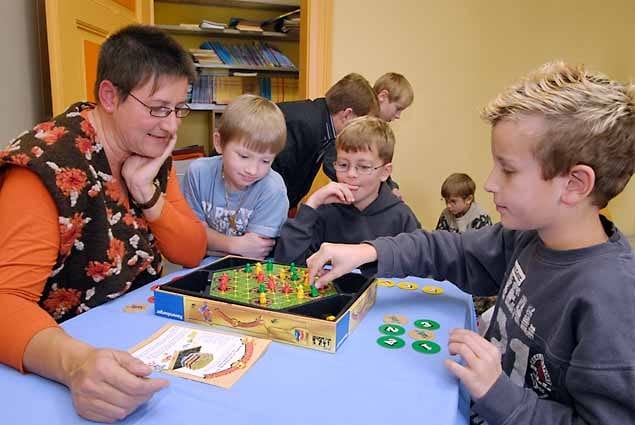 Farbiges, hölzernes Brettspiel am Tisch mit mehreren Kindern, Spielfiguren auf dem Spielplan und sichtbaren Spielkarten in einer hellen Gruppe